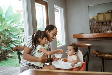 father and daughter feeding her baby sister together at home