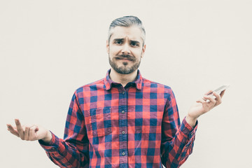 Doubtful man holding smartphone and looking at camera. Front view of uncertain bearded man in checkered shirt holding mobile phone and gesturing with hands. Technology concept