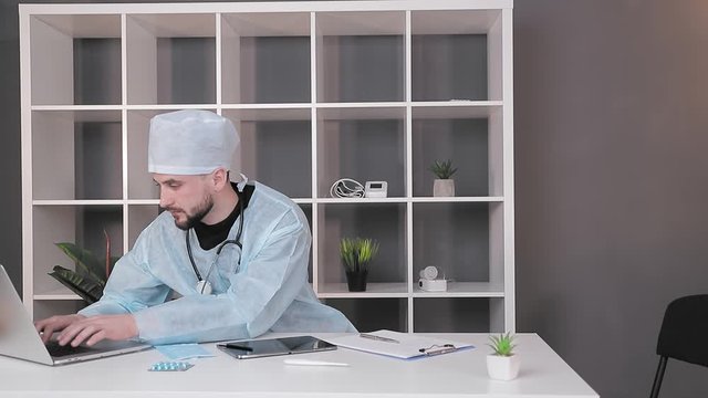 A doctor who works at the workplace in the office at the computer. A doctor in medical clothing is typing on a computer.