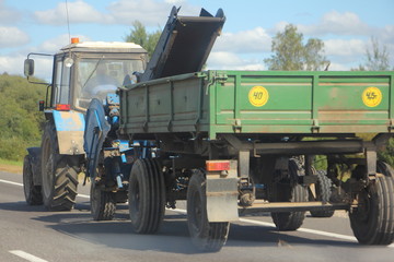 Fototapeta premium Tractor with flatbed trailer rides on a country road on a Sunny summer day, agricultural mechanization, rear side view in perspective