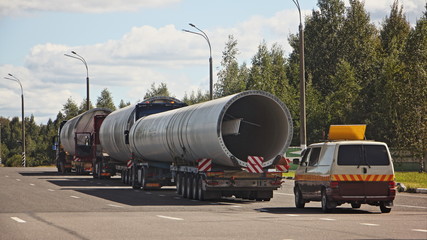 Long oversized cargo transportation logistics, big large pipes on 5-axle lowbed trawls semi-trucks with an escort car on highway at summer day