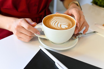 the hands of a girl who holds a coffee mug with a beautiful foam and next to it lies a diary and pen