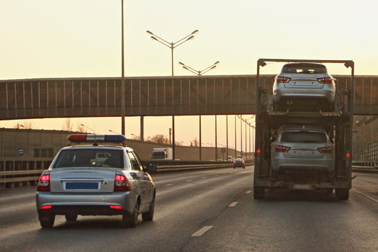Two-level russian car carrier rides on the highway in front of a police car in the evening against background of a pedestrian bridge, large-size road transport with a height restriction of the cargo