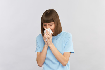 young woman in blue shirt blows her nose into white tissue isolated on grey