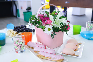 on a white dining table is a pink box with flowers of chrysanthemum, roses and lilies, next to it are eclairs, sweets in a glass and cake