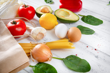 fresh vegetables and fruit spilled from a paper grocery bag on a white wooden background, peppers, tomatoes, avocado, lettuce, lemon, pasta, eggs, garlic, onions, spinach, detox;  rustic;