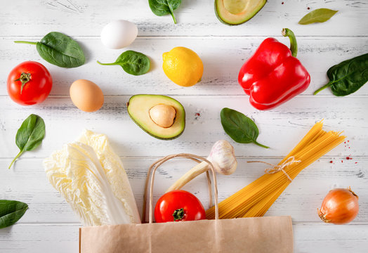 fresh vegetables and fruit spilled from a paper grocery bag on a white wooden background, peppers, tomatoes, avocado, lettuce, lemon, pasta, eggs, garlic,  spinach, detox; bio; rustic;
