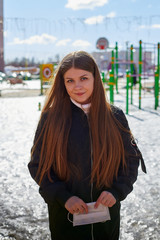 Beautiful young woman with long hair in spring clothes on the street with white medical mask on. Protect of girl against infection with coronavirus Covid19 in Russia in russian city