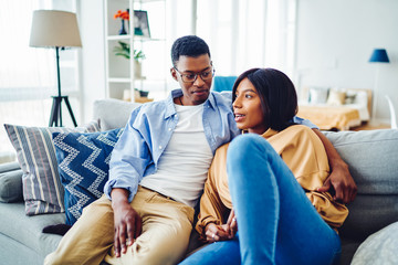 Carefree dark skinned marriage enjoying home recreation during weekend in cozy apartment with loft interior, African American male and female 20s hugging at comfortable couch in living room