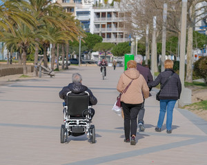 Obraz premium Group of adult friends walking along a promenade on sunny day. Elderly disabled person or senior on electric wheelchair or mobility scooter going with family. Modern technology for people disabilities