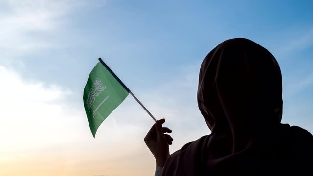 Time lapse. lonely Muslim woman in scarf with Saudi Arabia swaying flag on sunset sky. 