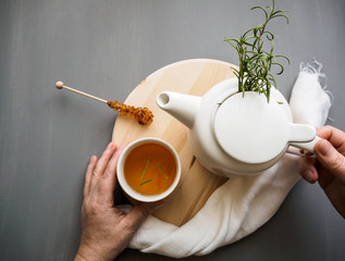 Hand mit einer Tasse Kräutertee und eine Teekanne auf einem Serviertablett aus Holz, grauer...