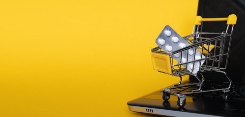 Grocery cart with pills on a black laptop on yellow background: place for text, banner, delivery of necessary medicinesduring quarantine, epidemics, diseases, assistance. Delivery, online shopping