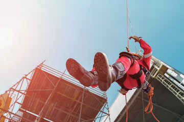 Close up pic of male rope access jobs worker wearing equipment safety full harness, working at height abseiling down from the high-rise construction site on structure steel background.