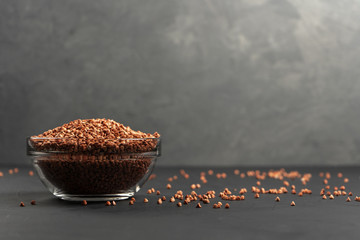 Buckwheat in a glass bowl near the copy space with spilled cereals on a black background.