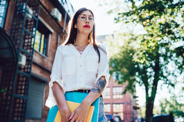 Half length portrait of attractive female designer standing at urban setting of sunny town and holding education sketchbook and notepad for creating content, Hispanic hipster girl in optical eyewear