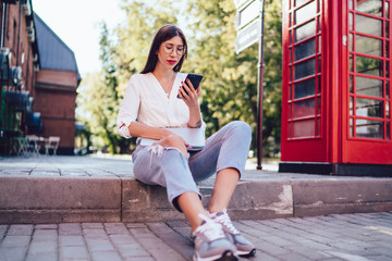 Attractive millennial woman in casual apparel resting near red telephone using cellphone gadget for online communication with followers, charming female blogger installing new app on smartphone