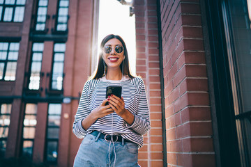 Half length portrait of cheerful meloman dressed in stylish apparel enjoying music songs from audio podcast playlist on modern cellphone device, happy woman in sunglasses listening stereo record