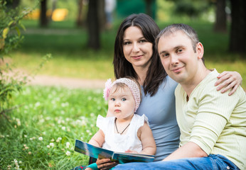 Obraz premium Happy young parents sitting in park with their small baby daughter on knees on summer clear day. Happy family time together concept
