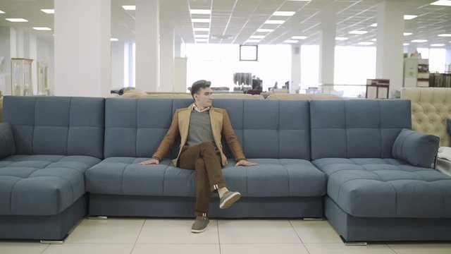 Man Sitting On Sofa At Furniture Store. Young Man Choosing Furniture For Home In Shopping Center. Customer Trying New Sofa And Touching Sofa Textile Upholstery For Quality.