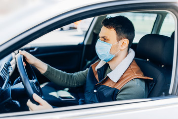 Beautiful young masked man sitting in a car, protective mask against coronavirus, driver on a city street during a coronavirus outbreak