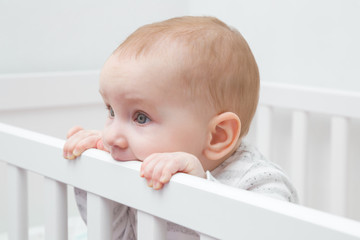Baby girl chewing edge of white crib because growing first tooth. Closeup.