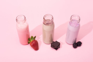 High angle view of bottles of milkshakes with strawberry, blackberries and pieces of chocolate on pink background
