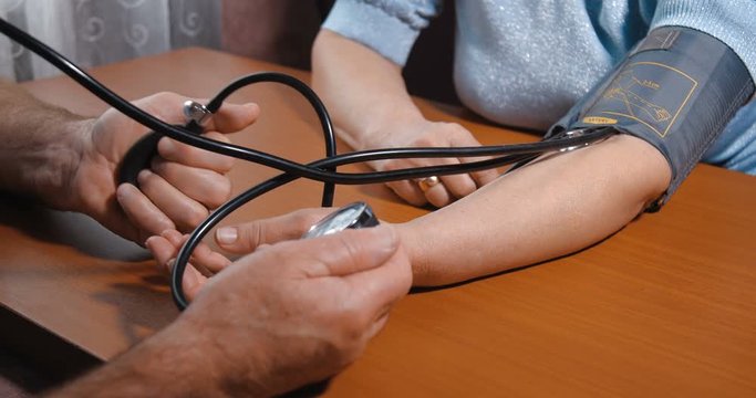 Close-up detail shot of hands of an elderly couple. Husband takes care of his wife by measuring her blood pressure using a mechanical tonometer on a brown wooden table at home. 4k 50fps slow motion