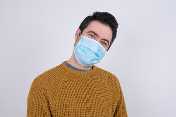 Shy blond boy wearing medical face mask smiling looking to the camera. Caucasian charming young man sanding against gray studio background feeling shy.