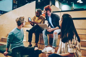 Dark skinned hipster girl holding mobile phone showing app to crew of colleagues on brainstorming session, positive skilled group of designers having meeting using modern technology for research