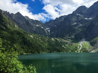 lake morskoe oko in the Tatra mountains with clear blue and green water, snow on the mountain tops and bright greenery of growing trees and bushes. Mountain landscape in the rays of the Golden sun.