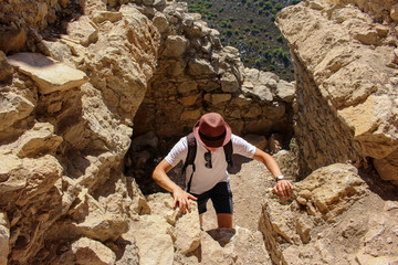 A young sportsman-tourist in a hat travels with a backpack through the mountains and climbs higher and higher on the ruins of an ancient castle between large blocks of stone against a clear sky.