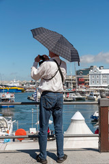 Cape Town, South Africa. 2019. Man holding an umbrella as sunshade and using a camera to take a picture of the waterfront.