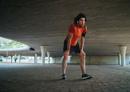 Attractive sporty young man in runner sportswear breathing gasping and taking a break after running workout under the bridge - man running in the new year