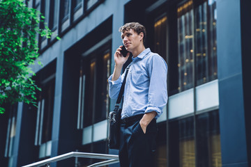 Caucasian owner of business calling to operator for online consultancy via cellular gadget connected to roaming outdoors, handsome male talking via modern device standing near enterprise corporation
