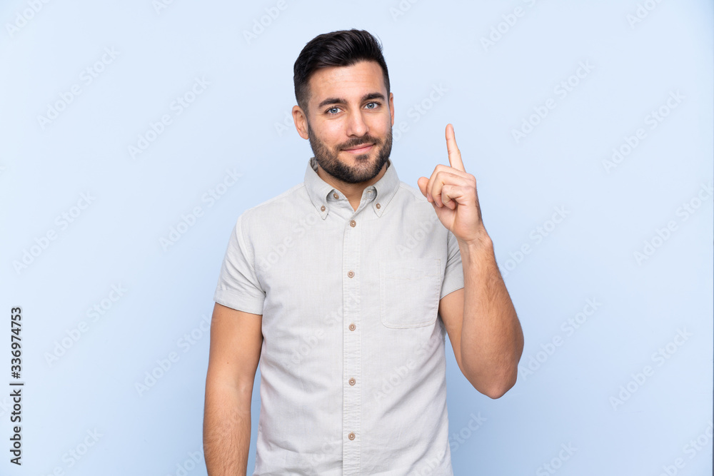 Wall mural young handsome man with beard over isolated blue background pointing with the index finger a great i