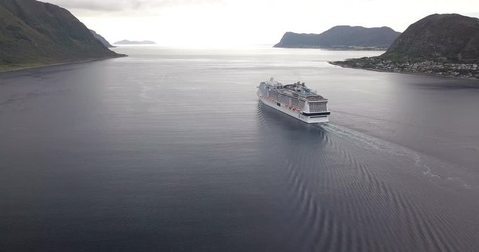 Panoramic and cinematic descending aerial drone hyperlapse of a luxurious cruise ship that is leaving a fjord towards the open sea in Norway at sunset.