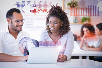 Happy colleagues working together and virtual statistic graphics. Cheerful man and woman sitting at table in office, using laptop, looking at screen, smiling, talking. Teamwork concept