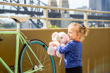 little girl with blond hair, holding a stuffed elephant in her hand, standing by a bicycle, in...