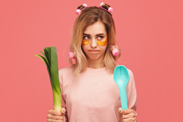 Girl purses lips, looks sideways, makes mistake, feels failure. Young woman with curlers in hair, patches on face (fabric mask under eyes for beauty), pajamas cooks stands on isolated pink wall Studio