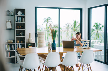 Cheerful blonde female freelancer enjoying remote job in comfortable home interior using modern gadgets, happy 20s woman blogger digital nomad satisfied with online income and media project