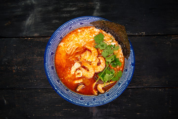 Top view on appetizing asian japanese tom yam ramen noodle soup with shrimps and squid in a traditional blue bowl on the dark wooden table, horizontal