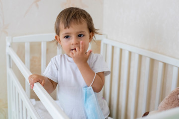 quarantine coronavirus pandemic concept. little cute toddler is safe in the baby cot at home and holds a medical mask.close up, copy space.