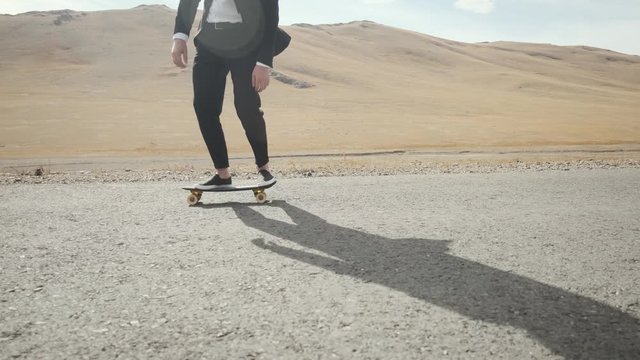 Caucasian man in black suit with tie and white shirt, an office worker happy excited man walks with skateboard along an empty scenic mountain road Mongolia steppe 4k close up