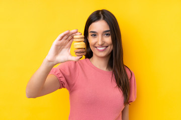 Young brunette woman over isolated yellow background holding colorful French macarons and with happy expression