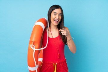 Lifeguard woman over isolated blue background with lifeguard equipment and surprised expression while pointing front - obrazy, fototapety, plakaty