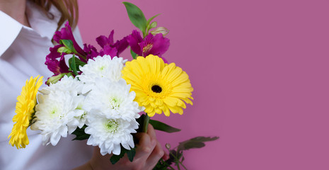 The girl on a pink background holds a beautiful bouquet of different flowers and covers her face.