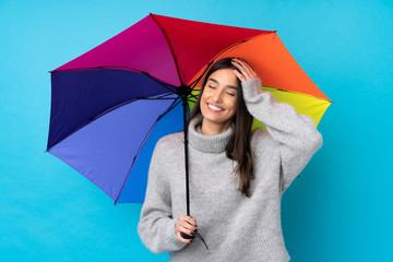 Young brunette woman holding an umbrella over isolated blue wall laughing