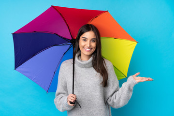 Young brunette woman holding an umbrella over isolated blue wall holding copyspace imaginary on the palm