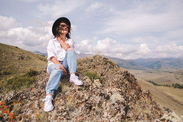 Naklejka premium Attractive brunette in a hat and white shirt is enjoying nature in the mountains. Beautiful young woman in a shirt and jeans walks along the beautiful landscape